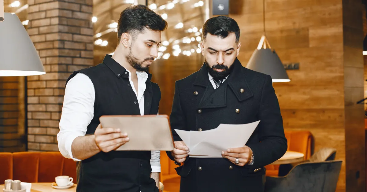 Two restaurant staff members converse inside a café/lounge area, reviewing figures on reports.