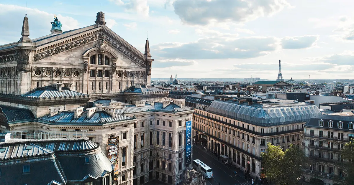 Wide-angle daytime view of the Palais Garnier Opera House and surrounding Haussmannian architecture in Paris under a clear blue sky, with the Eiffel Tower visible in the distant background.