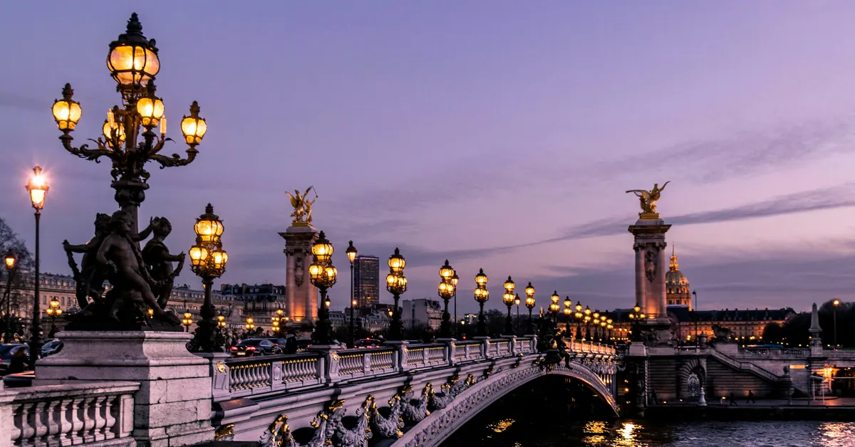 Pont Alexandre III in Paris at dusk, featuring ornate Art Nouveau lampposts glowing against a purple twilight sky, with the golden dome of Les Invalides and a distant skyscraper visible in the background.