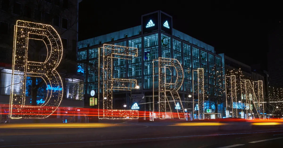 Large scale illuminated BERLIN letters at night on the Tauentzienstraße, featuring long-exposure light trails from city traffic against the backdrop of the Adidas flagship store and urban commercial architecture.