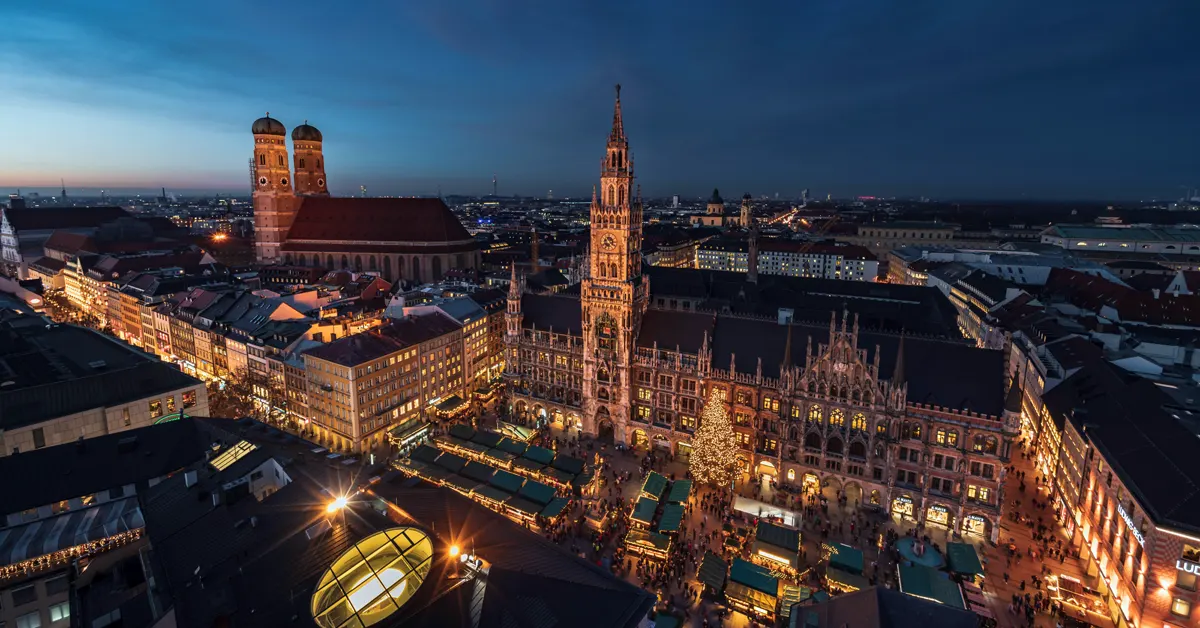 High-angle evening view of Marienplatz in Munich, Germany, featuring the illuminated New Town Hall (Neues Rathaus) and the twin towers of the Frauenkirche during a winter market.