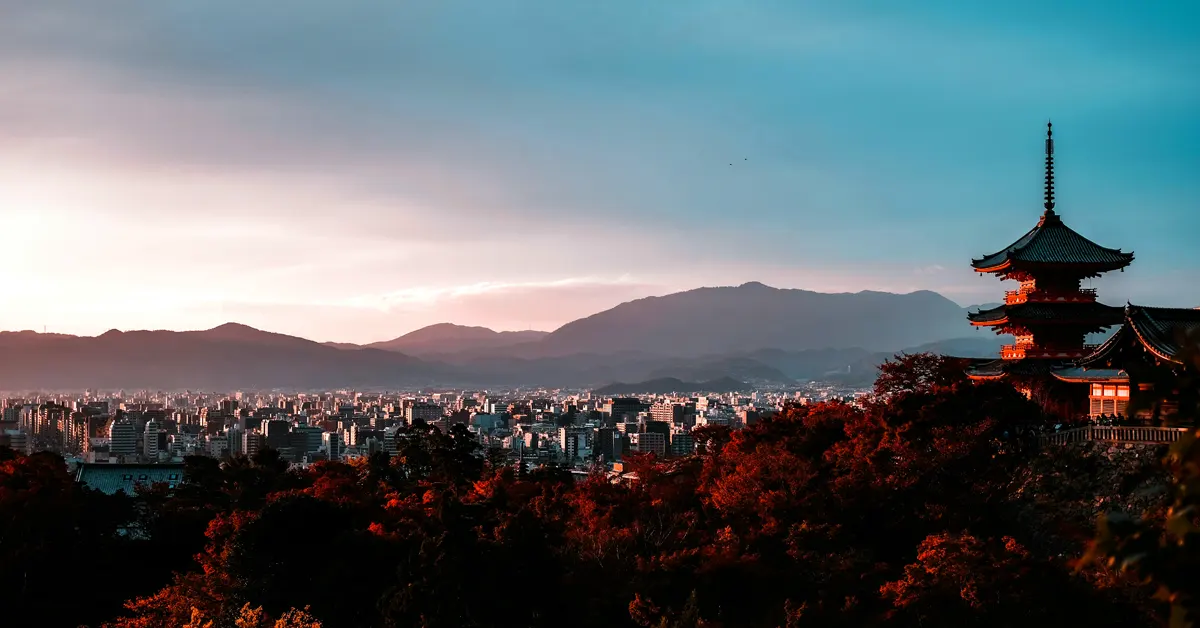 Panoramic twilight view of the Kyoto cityscape from an elevated vantage point, featuring the silhouette of a traditional multi-tiered pagoda on the right, surrounded by autumn foliage with mountains in the background.