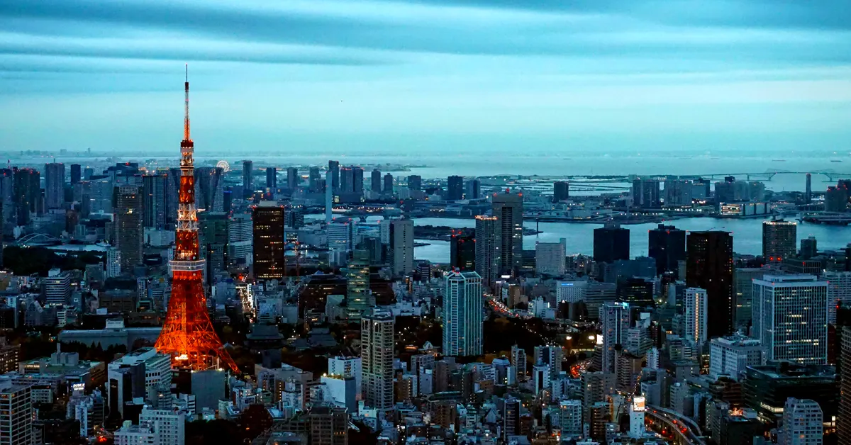 An elevated panoramic view of the Tokyo skyline at dusk, highlighting the illuminated Tokyo Tower in red and white against a backdrop of dense urban skyscrapers and the bay.