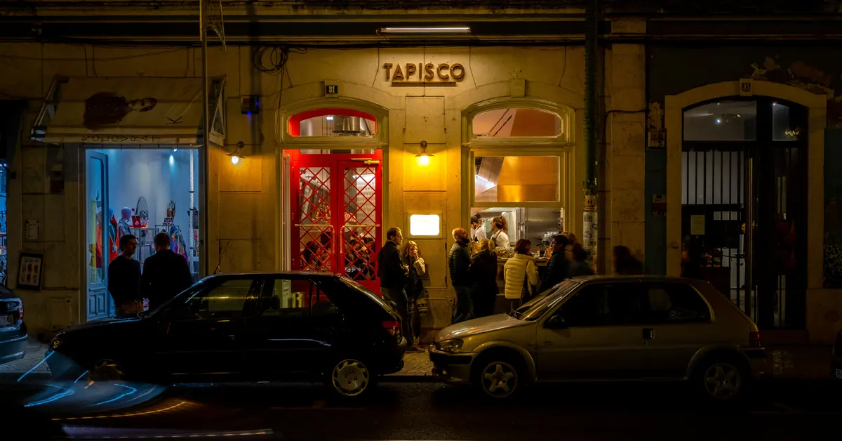 Alt Text Exterior night view of Tapisco restaurant in Lisbon, showing people gathered outside illuminated windows, a bright red front door, and two parked cars in the foreground.