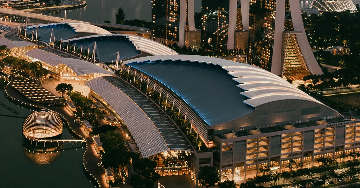 High-angle evening view of the Sands Expo & Convention Centre and The Shoppes at Marina Bay Sands in Singapore, featuring illuminated curved roofs alongside the waterfront and the floating Apple Marina Bay Sands globe.