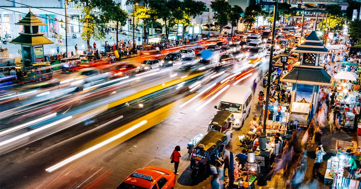 Long-exposure night view of a bustling, brightly lit street in Bangkok, Thailand, showing blurred light trails from heavy traffic, parked tuk-tuks, and vibrant sidewalk market stalls crowded with people.