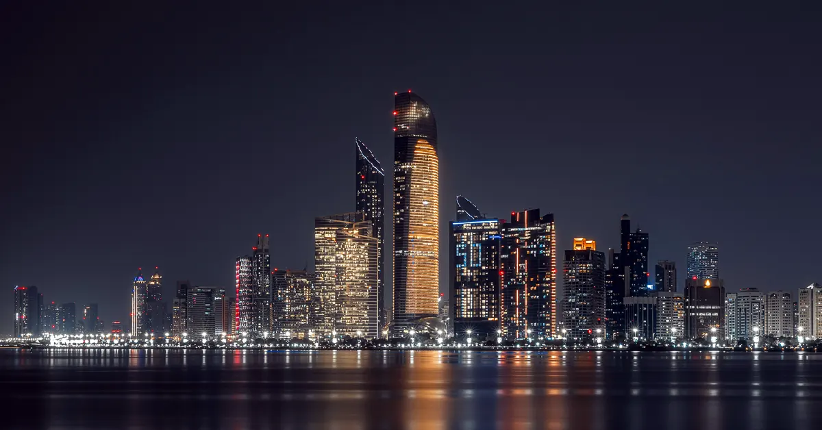 A panoramic night view of the Abu Dhabi skyline, United Arab Emirates, showcasing illuminated high-rise commercial and hospitality towers reflecting across the dark coastal waters.