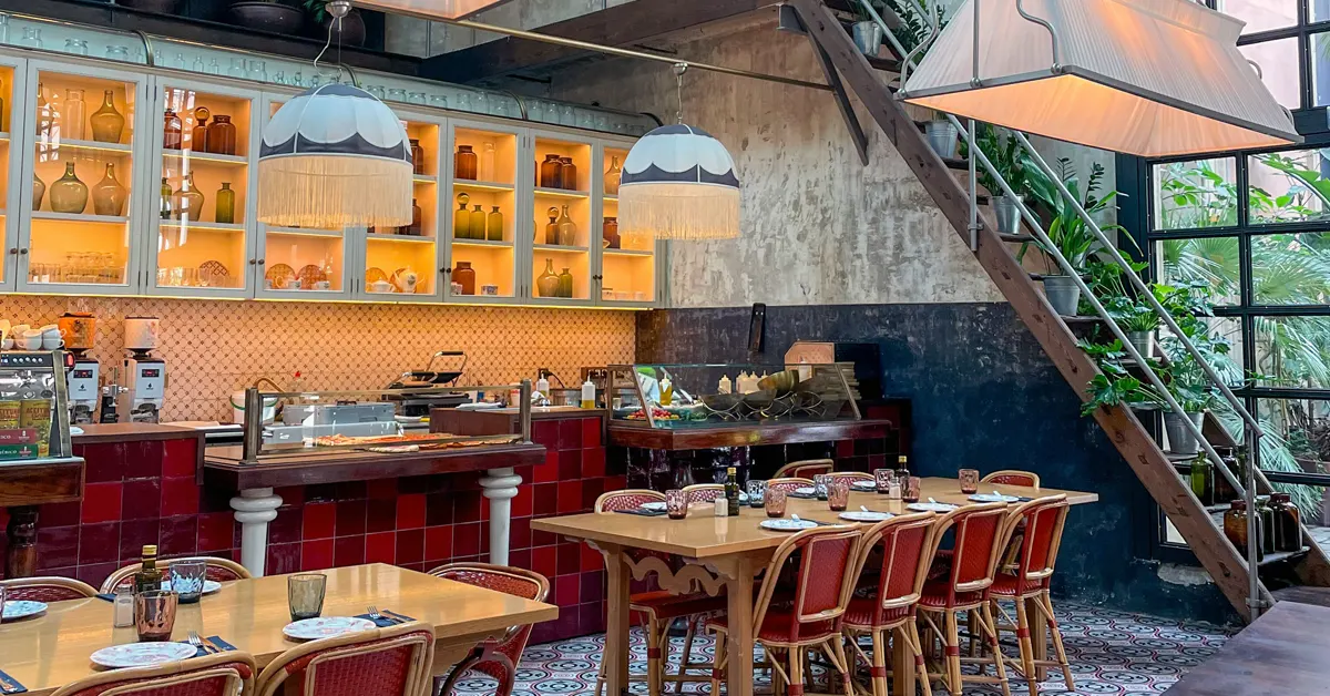 Vibrant interior of El Nacional restaurant in Barcelona, Spain, featuring set wooden dining tables with red-framed chairs, a tiled display counter, backlit bar shelving filled with glass bottles, decorative hanging lamps with fringes, a rustic wooden staircase with green potted plants, and patterned mosaic tile flooring.