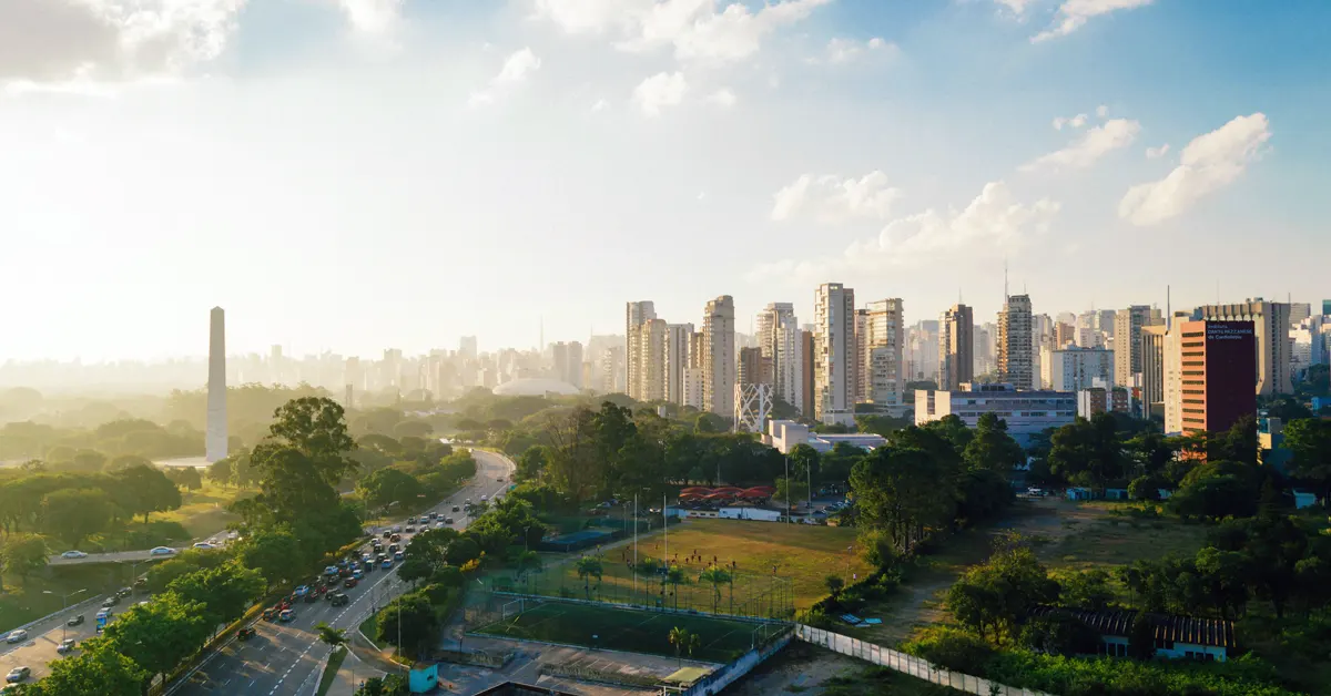 Aerial view of the São Paulo skyline with dense high-rise buildings, urban green spaces, and a busy highway under bright daylight.