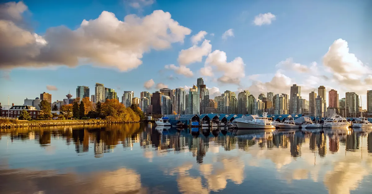 Panoramas of the Vancouver downtown skyline with high-rise buildings and skyscrapers reflected on the calm water of Coal Harbour, viewed from Stanley Park under a partly cloudy sky at sunset.