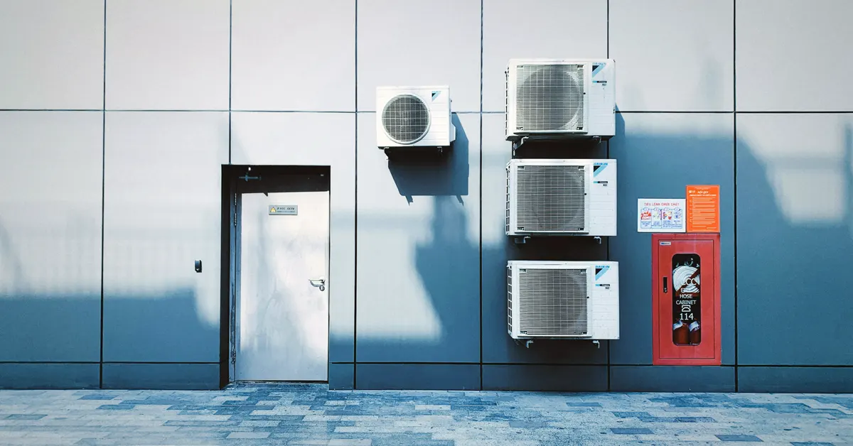 Four white commercial air conditioning units mounted on a sleek, grey paneled exterior wall next to a silver door and a red fire extinguisher cabinet.