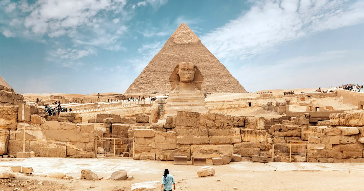 The Great Sphinx of Giza with the Pyramid of Khafre rising in the background under a blue sky with scattered clouds, viewed from a low angle with visitors visible in the distance.