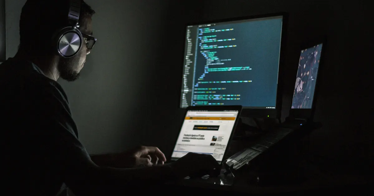 A person in a dimly lit room wearing headphones and glasses, looking at multiple computer screens displaying lines of code and websites, symbolizing online cyber fraud or account takeover hacking.