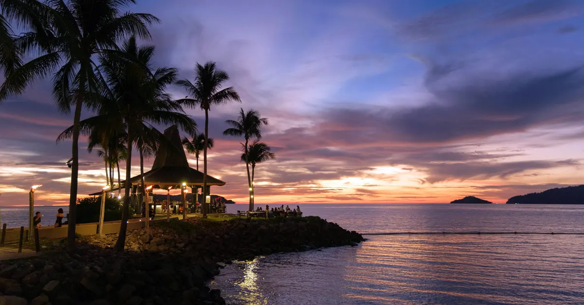 Silhouetted palm trees and an open-air pavilion bar on a rocky coast in Kota Kinabalu, Malaysia, under a vibrant purple, orange, and blue sunset sky overlooking the sea.