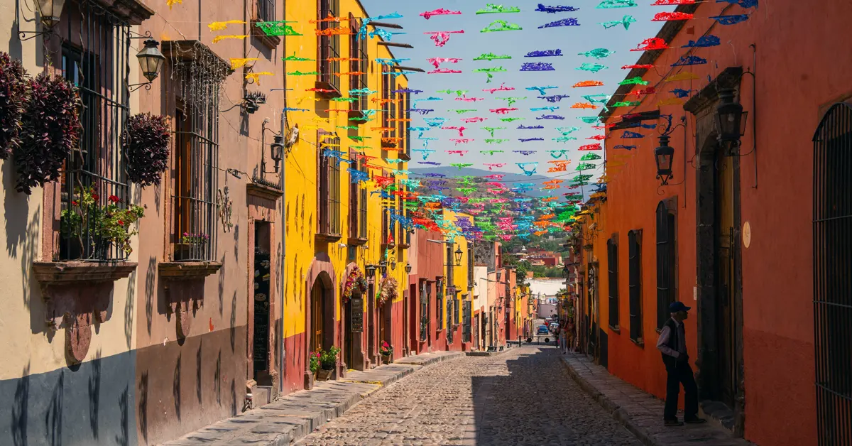 A narrow, sloping cobblestone street in Mexico lined with vibrant yellow, orange, and terracotta colonial-style buildings under a clear blue sky. Strands of multi-colored papel picado banners hang overhead across the street, creating shadows on the stone road. A single person stands in the shade on the right side of the street.