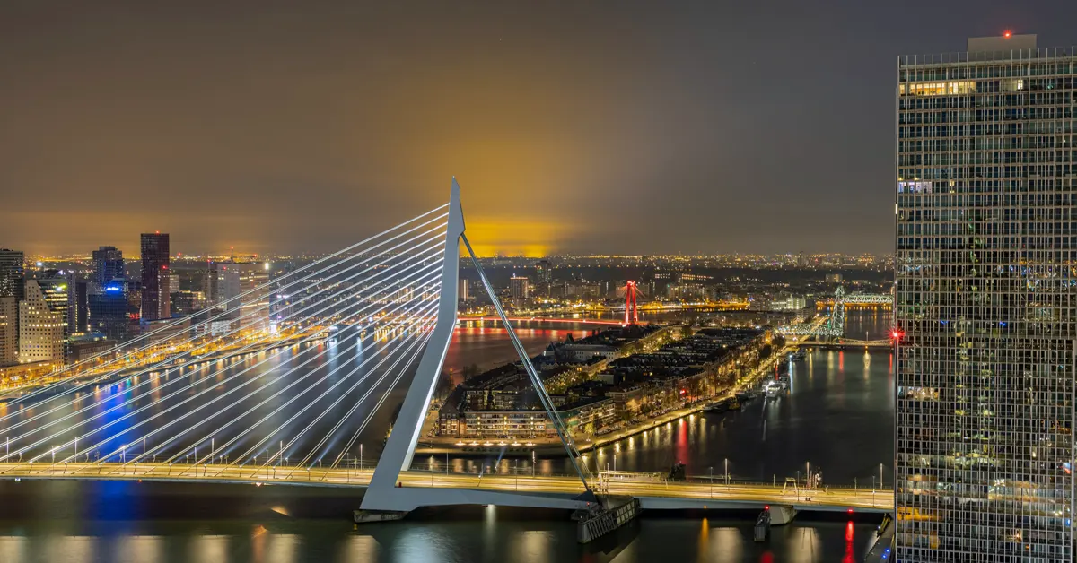 Nighttime aerial view of the illuminated Erasmus Bridge and urban skyline in Rotterdam, Netherlands, featuring modern architecture along the Nieuwe Maas river.