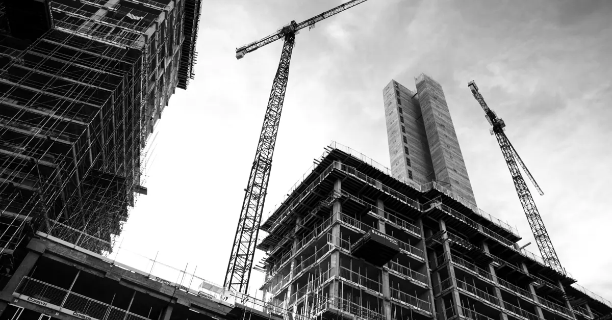 Low-angle black and white shot of modern high-rise buildings under construction with tower cranes against a cloudy sky.