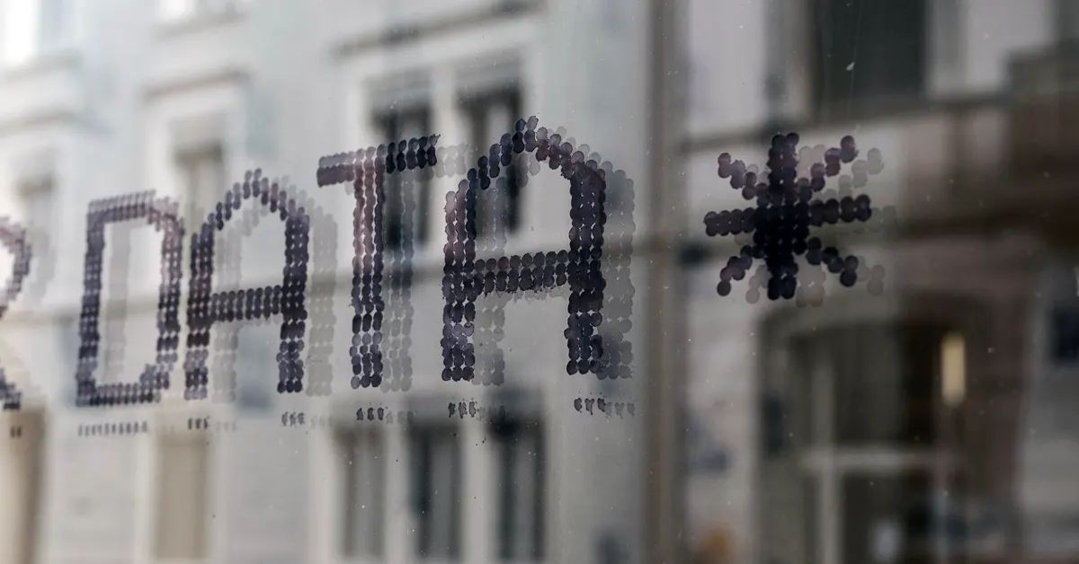 The word "DATA" written in a stylized, dotted font on a glass window pane, with a blurred building visible in the background, symbolizing data exposure and transparency.