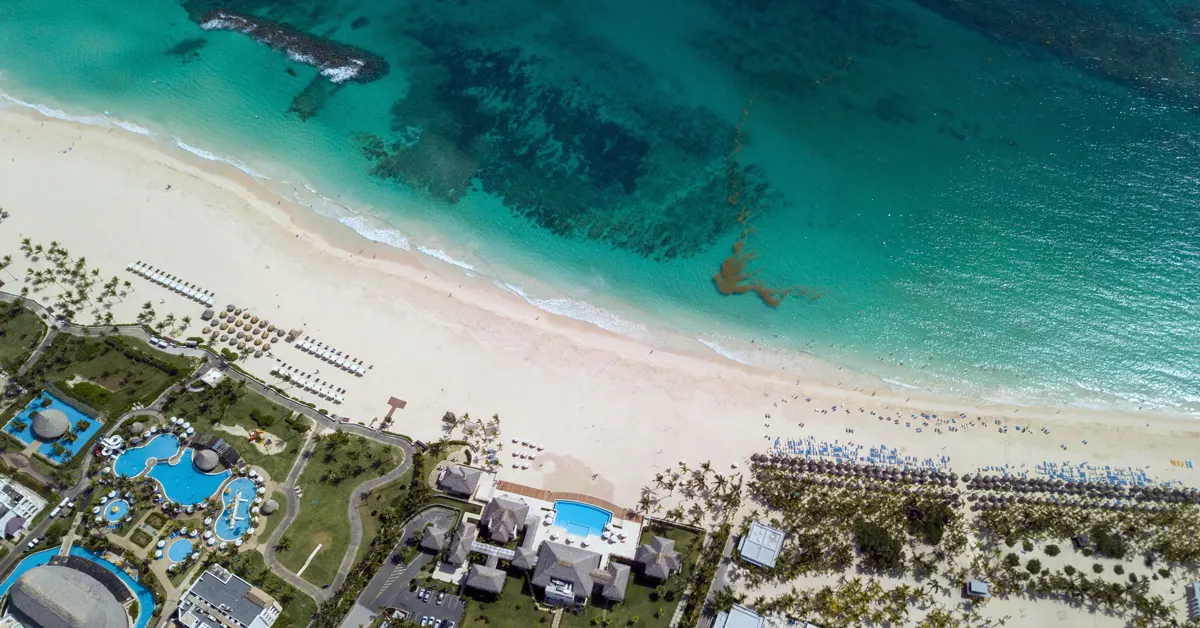 High-angle aerial view of a luxury beachfront resort in the Dominican Republic, featuring white sand beaches, clear turquoise ocean waters, organized rows of beach loungers, and expansive resort swimming pools surrounded by tropical landscaping.