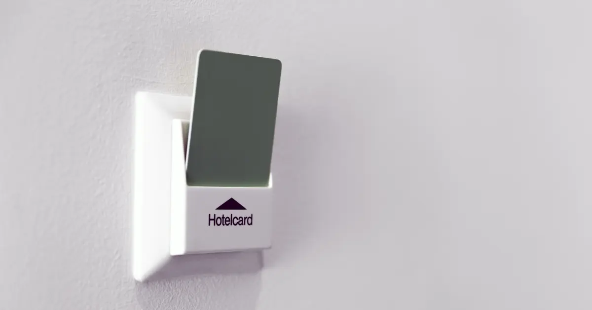 Close-up of a plain gray hotel key card inserted vertically into a white plastic wall-mounted power switch labeled "Hotelcard" against a plain white wall.