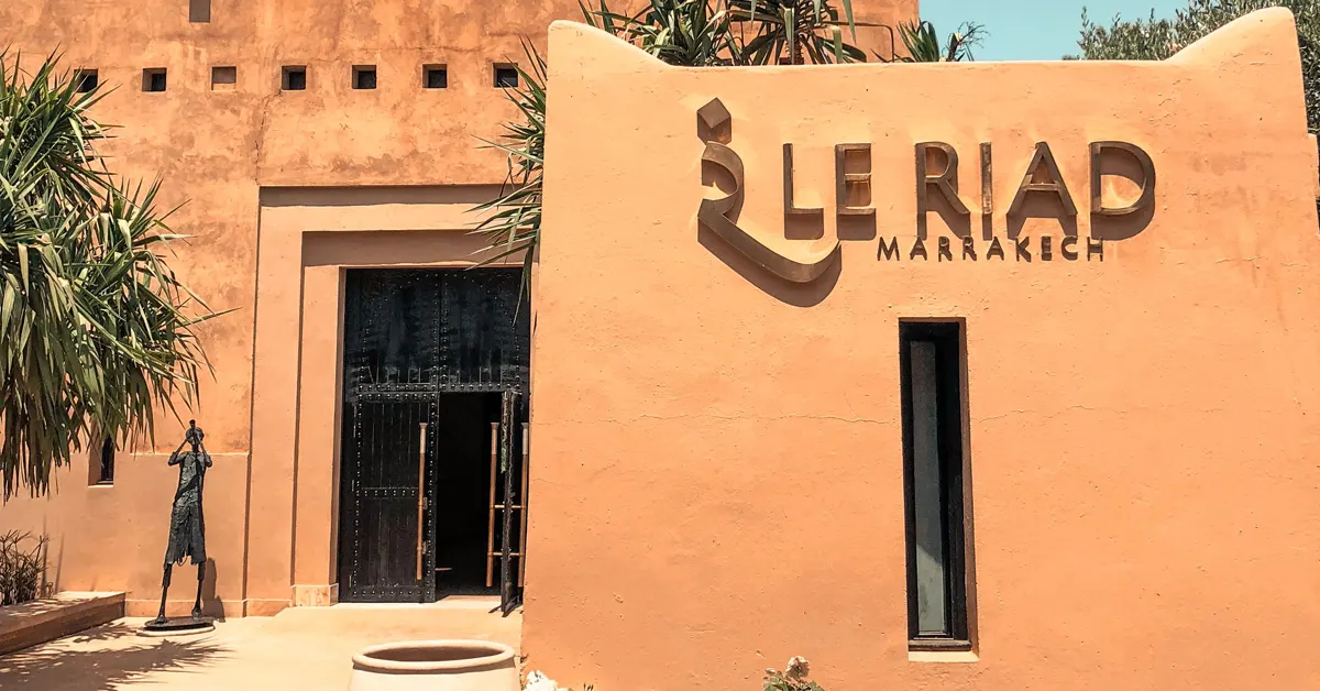 Exterior view of Le Riad Marrakech featuring terracotta-colored walls, a traditional dark wooden entrance door, and a modern metallic sign reading LE RIAD MARRAKECH.