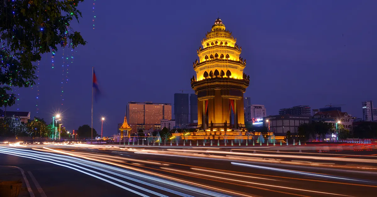 Illuminated Independence Monument in Phnom Penh at dusk with long-exposure traffic light trails in the foreground.