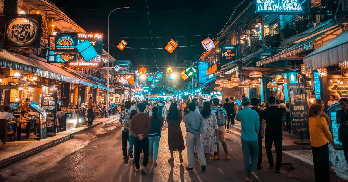 Crowds of pedestrians walk down Pub Street in Siem Reap at night, surrounded by illuminated neon signs, glowing lanterns, and open-air restaurants.