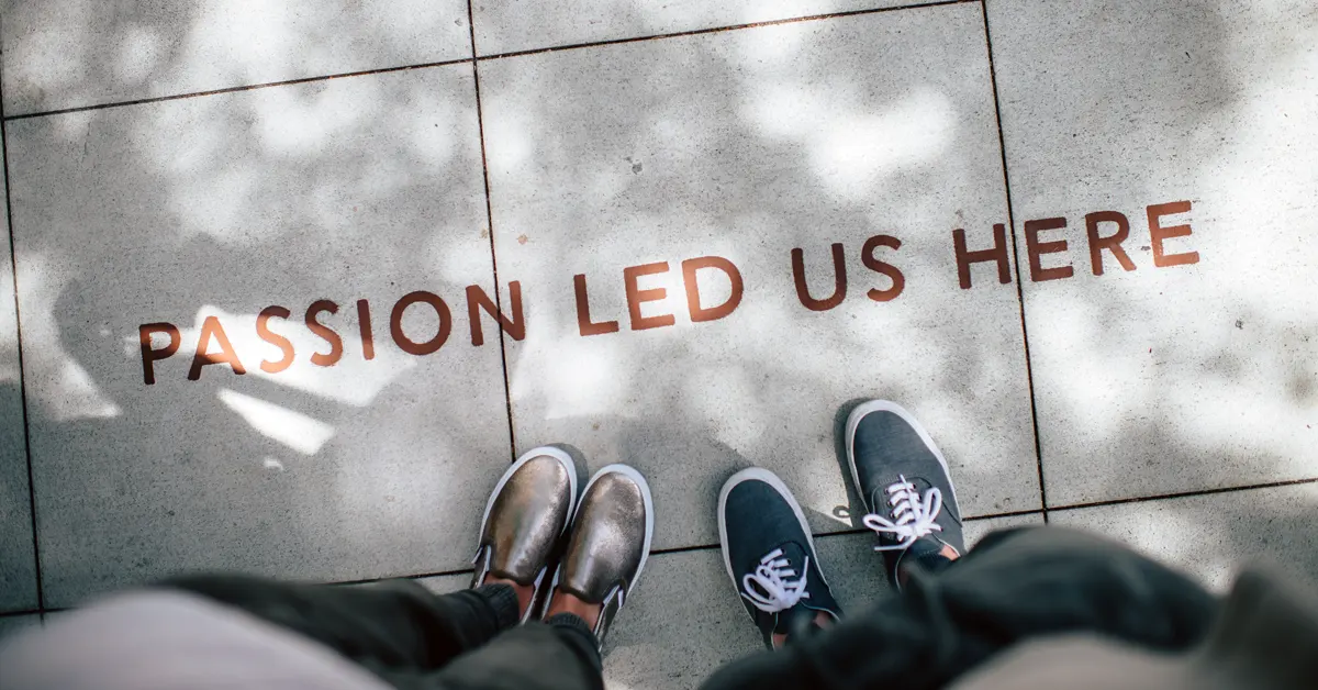 An overhead view of two sets of feet in casual shoes, one silver slip-on and one grey lace-up, standing on a light-grey concrete tiled sidewalk in front of the rose gold text 'PASSION LED US HERE'. Sunlight and dappled tree shadows are cast across the pavement.