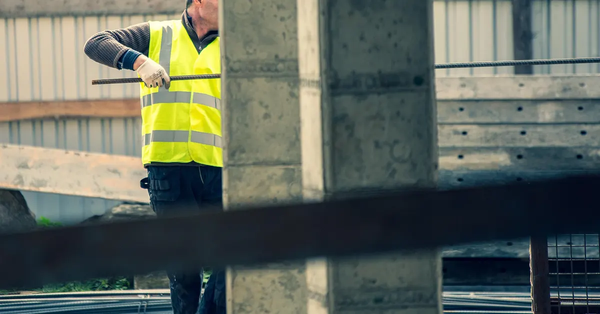 Skilled construction worker in a high-visibility safety vest carrying steel rebar on an active hotel development site.