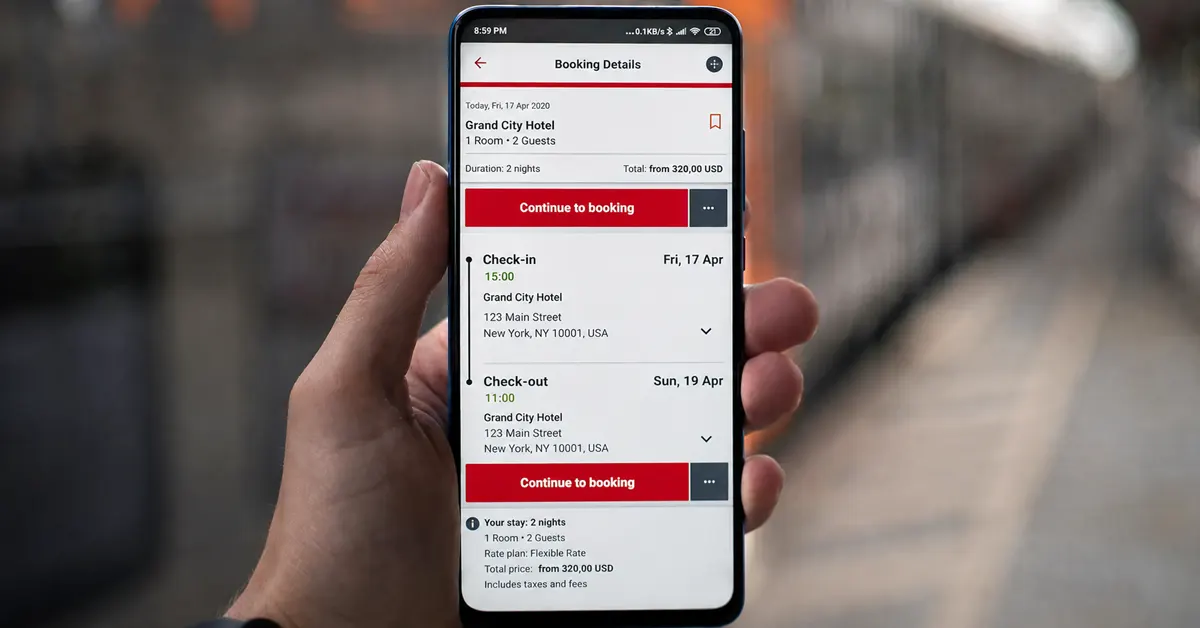 A hand holding a smartphone showing a hotel booking screen for Grand City Hotel, with a blurred train platform in the background.