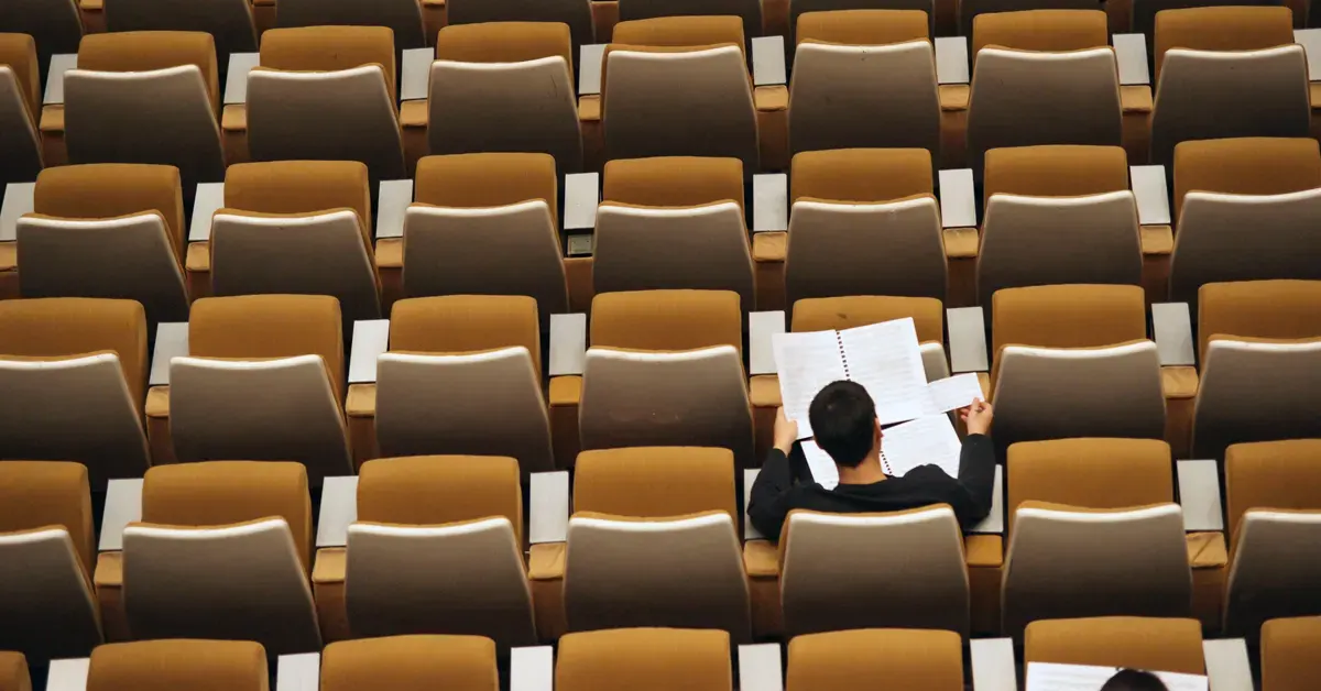 Overhead view of a single student seated alone in a large auditorium surrounded by empty brown chairs