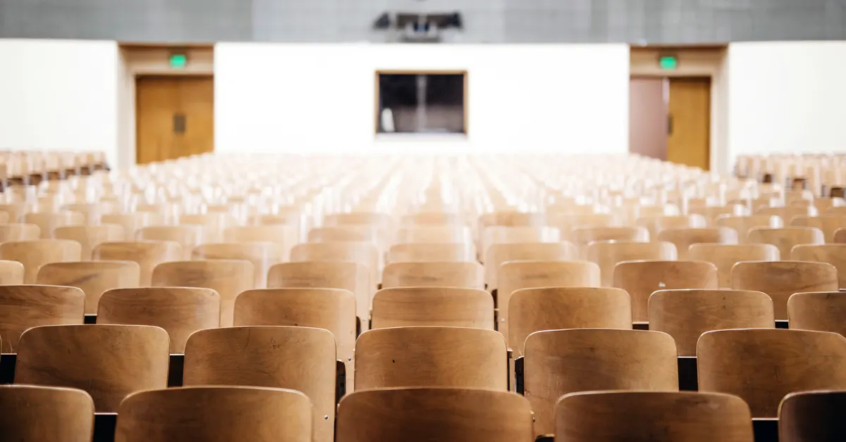 Rows of vacant wooden chairs facing an empty stage in a large, silent lecture hall