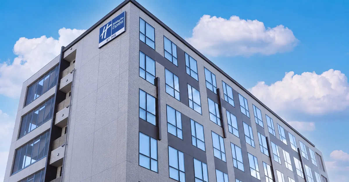 Low-angle view of a modern Holiday Inn Express hotel building with tiled facade and brand signage against a blue sky, part of IHG's portfolio conversion deal in Kyoto, Japan.