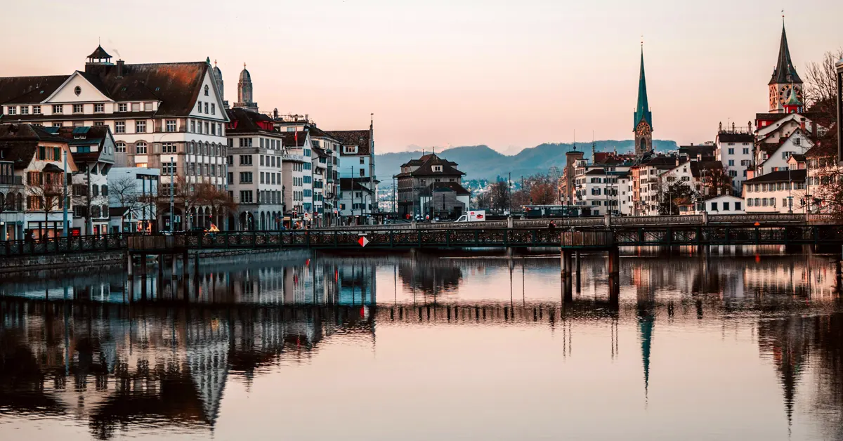 Wide view of Zurich's historic city center buildings and church spires reflected in the still waters of the Limmat River under a soft evening sky.