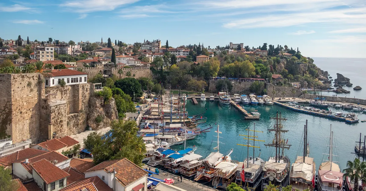 High-angle aerial view of Kaleiçi Old Town harbour in Antalya, Türkiye, showing traditional tour boats docked in turquoise water surrounded by historic stone walls, red-tiled roofs, and coastal cliffs.