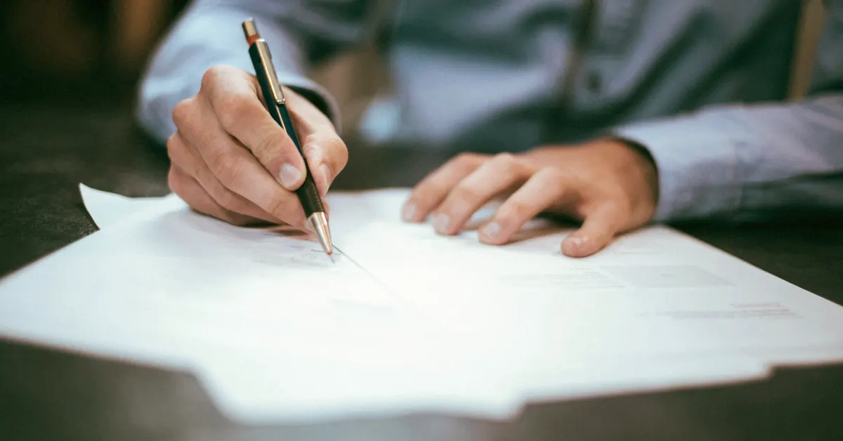 Close-up view of a person's hands holding a metallic mechanical pen and signing a paper document on a dark table.