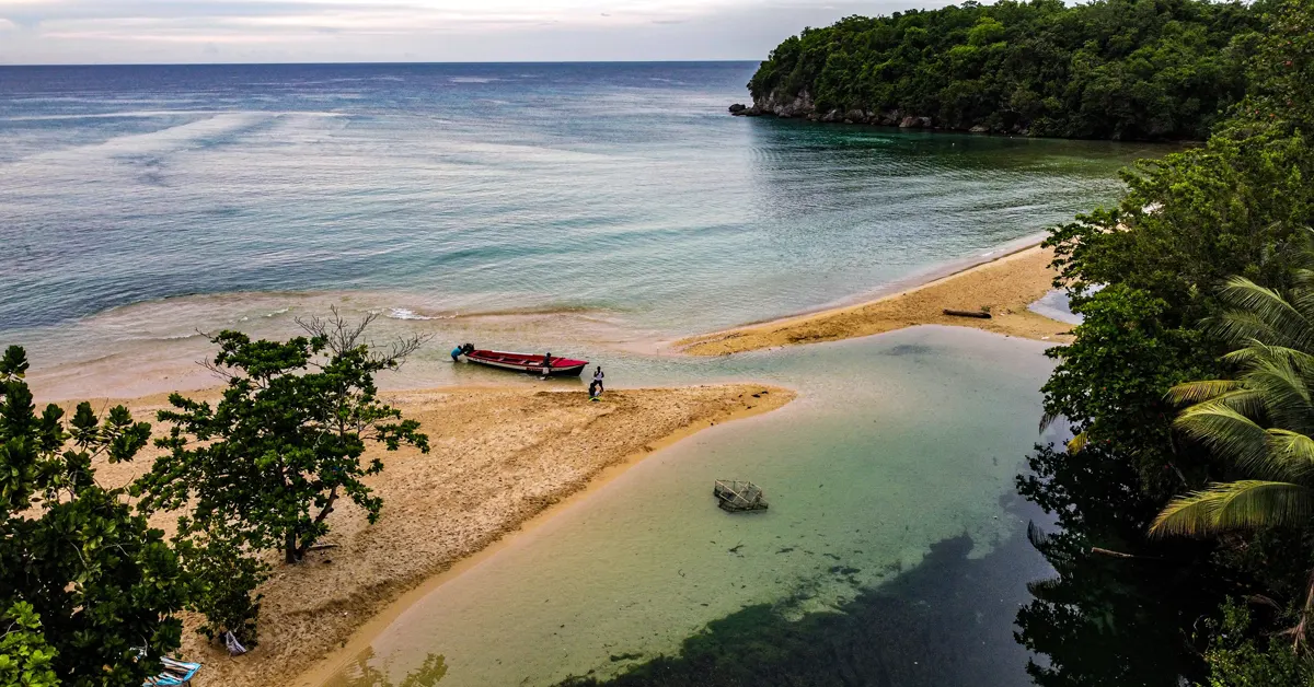 High-angle view of a sandy beach in Jamaica with a red and white boat anchored at the shallow water's edge near lush green trees.