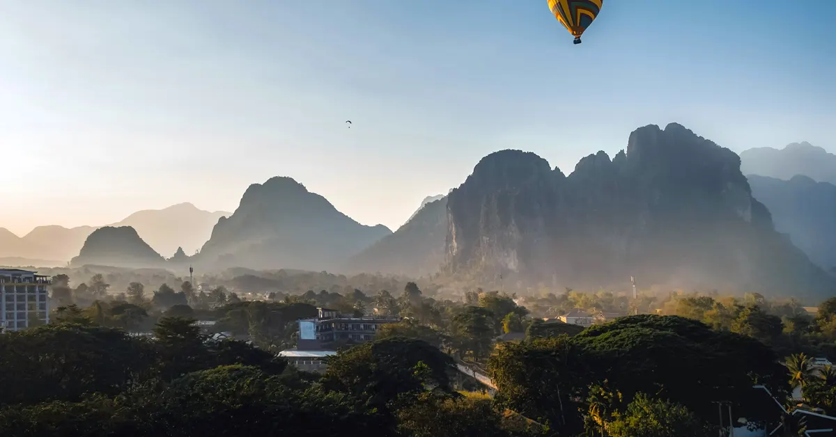 A colorful hot air balloon floats in a bright blue sky over dramatic limestone karst mountains and the hazy town landscape of Vang Vieng, Laos.