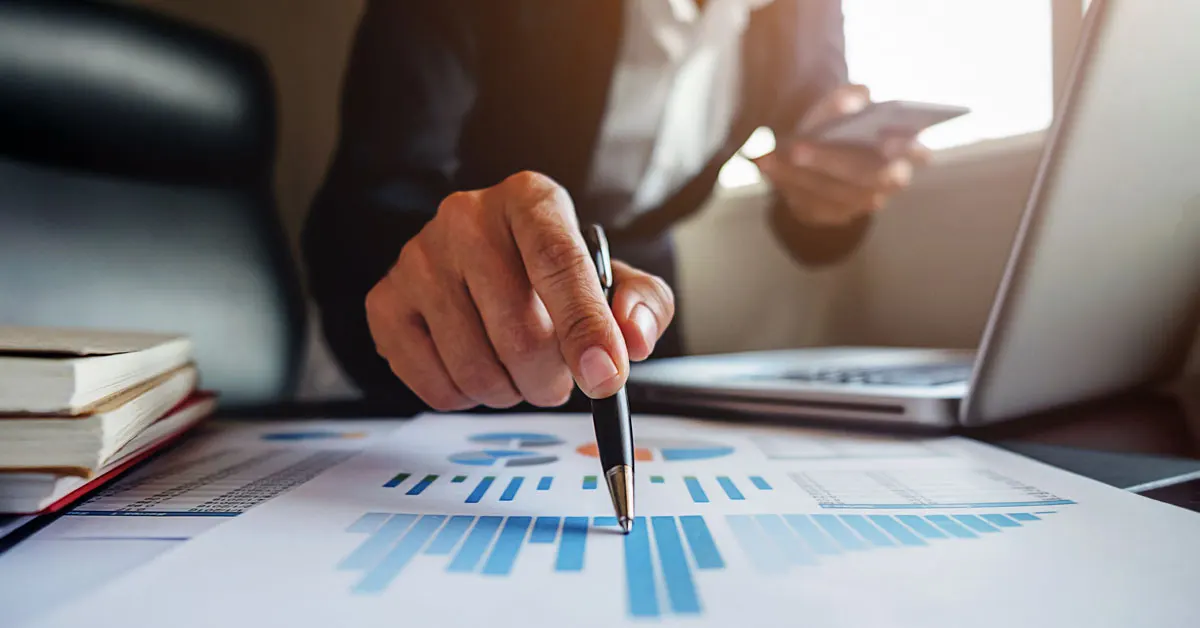 Close-up of a business professional in a suit pointing a pen at printed bar charts on a desk alongside a laptop and smartphone.