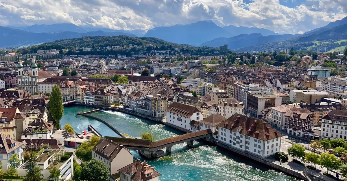 High-angle panoramic view of Lucerne, Switzerland, showing the Reuss River, historic architecture, the Spreuer Bridge, and distant mountain peaks under a partly cloudy sky.