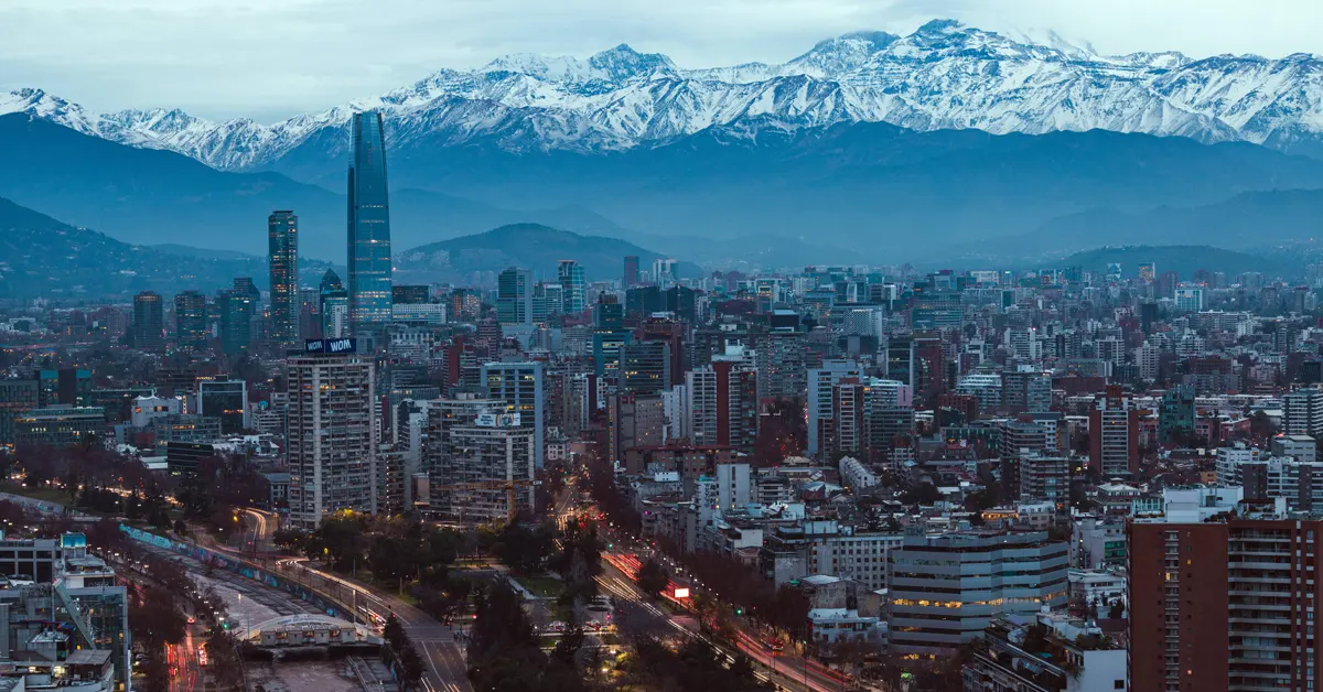 Panoramic view of Santiago, Chile's modern city skyline featuring high-rise buildings and the Gran Torre Santiago, with snow-capped Andes mountains towering in the background.