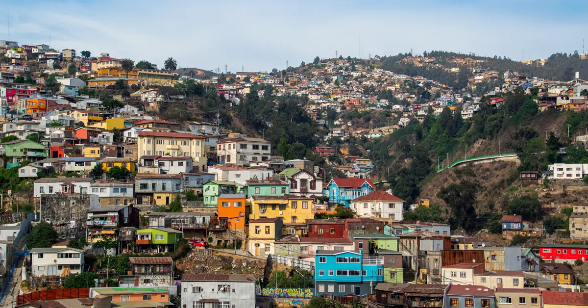 A wide, elevated view of brightly painted, multi-colored houses stacked densely along steep hillsides in Valparaíso, Chile under a clear sky.