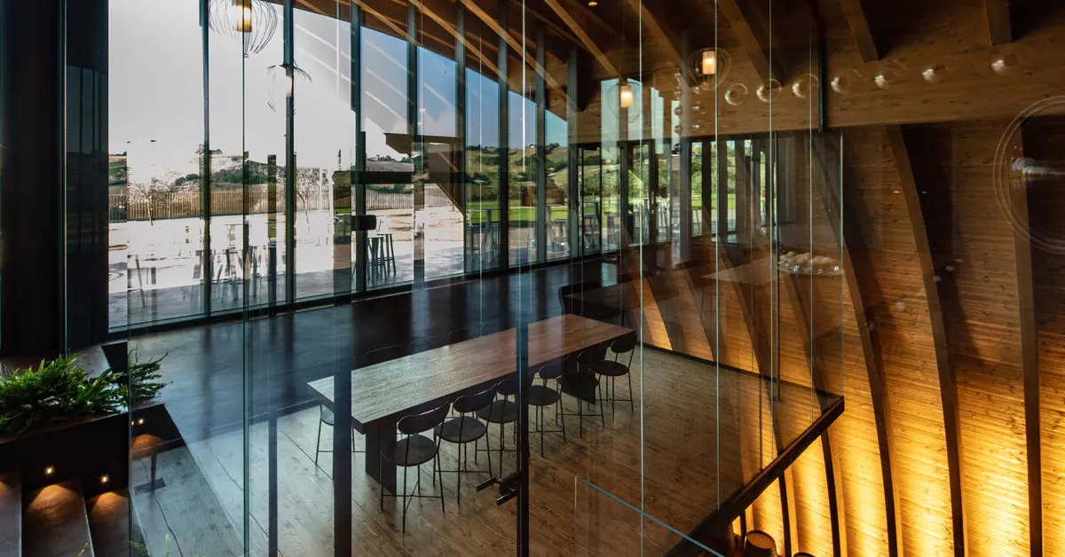 High-angle shot looking through a glass partition into a modern interior space featuring a long wooden conference or dining table surrounded by black chairs.
