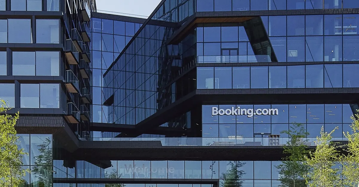 Modern dark glass building facade featuring the white Booking.com logo on a large window panel, with a connecting skybridge and green tree foliage at the base.